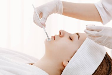 Rahat bir kadın parlak bir kaplıcada hafif bir yüz bakımı alır. Eldivenli bir estetikçi ağız etrafına bir fırça ile yatıştırıcı bir maske takarak sakin, dikkatli ve profesyonel bir cilt bakımı uygular..