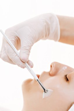Profesyonel bir yüz tedavisinde eldivenli bir estetisyen, parlak bir spa ortamında, gevşemiş bir müşterinin yanağına yelpaze fırçasıyla maske takıyor..