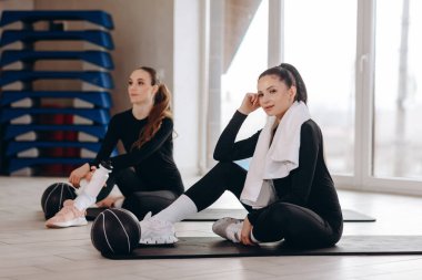 Siyah giysili iki genç kadın spor yaptıktan sonra paspasların üzerine oturur, sağlık, odak ve yoldaşlık sağlayan modern bir spor salonunda ilaç topları ve havlularla dinlenirler..