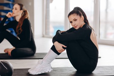 İki genç kadın antrenmandan sonra parlak bir spor salonunda paspasların üzerinde dinleniyor, siyah spor ayakkabılar ve beyaz spor ayakkabılar giyiyor, odaklanma, iyileşme, arkadaşlık ve sağlıklı aktif bir yaşam tarzı sergiliyorlar..