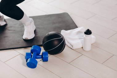 Mavi dambıllar, siyah bir ilaç topu, paspas ve spor ayakkabısının yanında havlu ve su şişesi, dayanıklılık eğitimi, hidrasyon ve aktif bir yaşam tarzı içeren ev çalışması sahnesi.