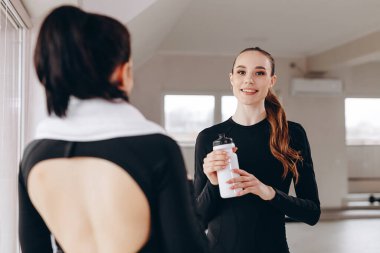 Spor giyim kuşamında iki kadın spor salonunda sohbet ediyor ve parlak bir spor salonunda gülümsüyor. Antrenmandan sonra su şişesini tutan biri gibi arkadaşlığı, zindeliği, susuzluğu ve antrenman alanında aktif yaşam tarzını aktarıyor..