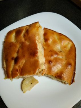 Beyaz bir tabakta iki dilim elmalı turta. Elmayı koyu bir yüzey üzerinde duruyor.