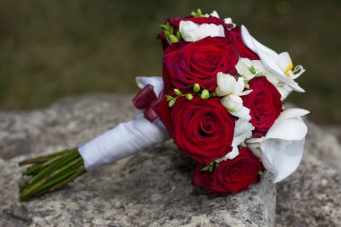 Kırmızı güller, beyaz frezeler ve orkidelerle süslü zarif bir gelin buketi, beyaz bir kurdeleyle bağlanmış ve bir kayaya yerleştirilmiş, aşkı ve kutlamayı simgeliyor.