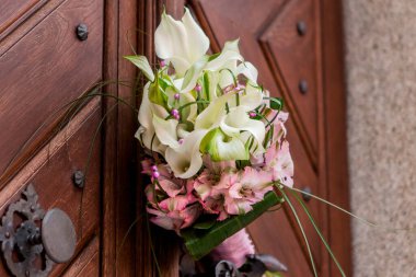 Beyaz zambaklardan ve pembe çiçeklerden oluşan güzel bir düğün buketi antik bir ahşap kapıya asılarak romantik ve zarif bir sahne yaratıyor.