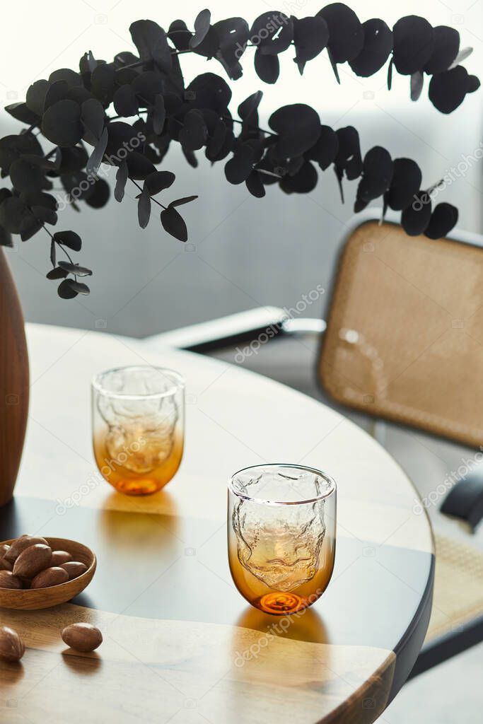 Stylish composition on the design table with elegant glasses, nuts and black eucalyptus flowers in wooden vase. Modern dining room interior. Template.