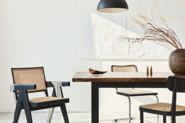 Ahşap masa, dizayn sandalyeleri, vazoda kurumuş çiçekler, siyah kolye lambası, duvarda resim resimleri ve zarif kişisel aksesuarlarla yemek odasının minimalist kompozisyonu. Şablon.