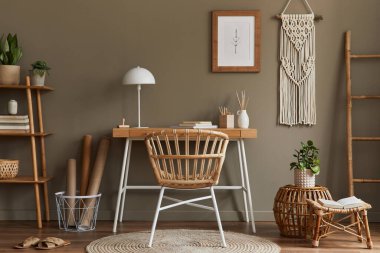 Ev ofis odasının içinde modern kompozisyon ahşap masa, şık koltuk, bambu raf, halı, makrome, poster çerçevesi, ofis malzemeleri, dekorasyon ve kişisel aksesuarlar..