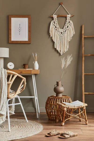 Stylish bohemian interior of home office space with wooden desk, rattan armchair, brown mock up poster frame, macrame, office supplies, lamp, decoration and elegant personal accessories in home decor.