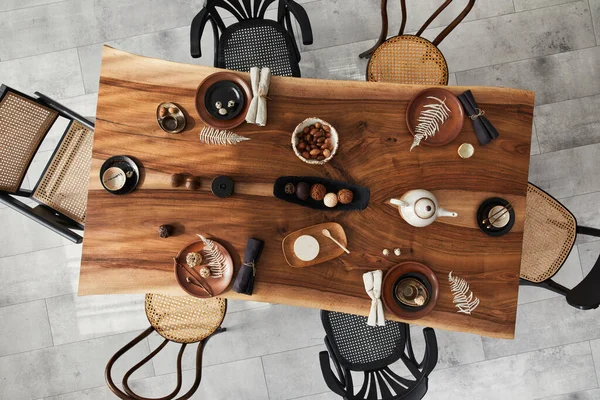 Stylish interior design of dining room with wooden walnut table, retro chairs, tableware, plates, tablecloth, teapot, food, decoration and elegant accessories. Cement floor. Template. Top view.