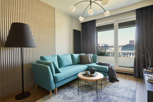 Creative composition of modern living room interior in small apartment. Eucalyptus sofa, pillows and plaid, modern designed chandelier and black lamp, small wooden coffee table and personal accessories. Big windows with big city view on. Carpet on th