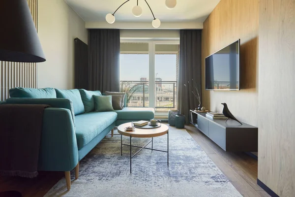 Creative composition of modern living room interior in small apartment. Eucalyptus sofa, pillows and plaid, modern designed chandelier, small wooden coffee table, black commode and personal accessories. Big windows with big city view on. Carpet on th