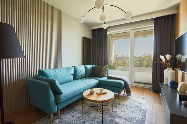 Creative composition of modern living room interior in small apartment. Eucalyptus sofa, pillows and plaid, modern designed chandelier, small wooden coffee table, black commode and personal accessories. Big windows with big city view on. Carpet on th