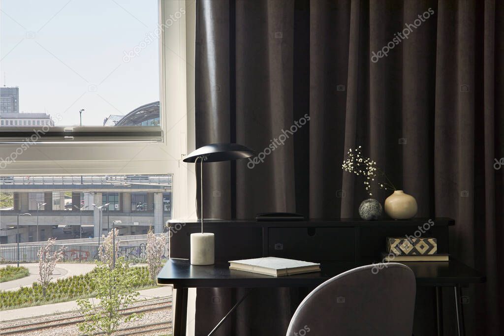 Stylish composition of small modern workspace interior with minimalistic lamp and personal accessories. Neutral walls with black panels. Panoramic windows with grey curtains. Minimalistic masculine concept. Template.