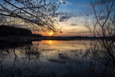 Nehir kıyılarından taştı. Su basmış topraklar ve çalılar. Batan güneş suya yansıyor. Önünde bir nehir ve dallar olan akşam manzarası.