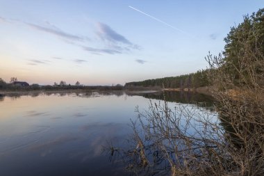 Nehirle akşam manzarası. Ufuktaki köy ve orman. Nehirde gün batımı. Gökyüzü ve akşam bulutları suya yansıyor..