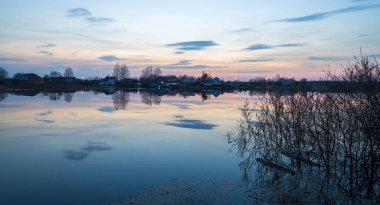 Nehirle akşam manzarası. Ufuktaki köy ve orman. Nehirde gün batımı. Gökyüzü ve akşam bulutları suya yansıyor. Ön planda ağaçların ve çalıların dalları.