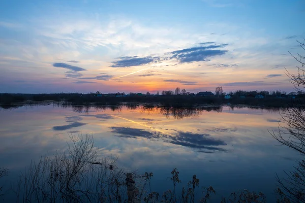 Evening landscape with a river. Village and forest on the horizon. Sunset on the river. The sky and evening clouds are reflected in the water.