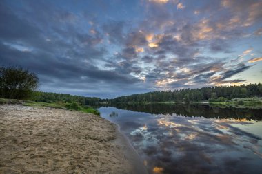 Akşamları nehir kıyısında yaz manzarası. Dramatik bulutlar suya yansıyor. Gün batımından sonra manzarada yoğun renkler.