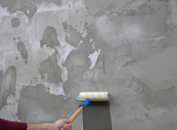 A worker applies the primer to the wall with a long-handled roller. A man\'s hand rolls a primer onto a gray wall. Wet footprint on a plastered wall. House facade renovation concept.
