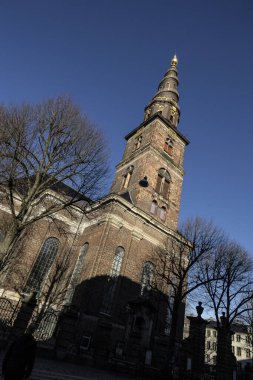 Danimarka, Kopenhag 'daki Kurtarıcımız Kilisesi' nin göz kamaştırıcı mimarisinin çıplak dallarla çerçevelenmiş görüntüsü kuleyi parlak mavi gökyüzüne doğru aydınlatıyor..