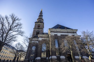 Danimarka, Kopenhag 'daki Kurtarıcımız Kilisesi' nin göz kamaştırıcı mimarisinin çıplak dallarla çerçevelenmiş görüntüsü kuleyi parlak mavi gökyüzüne doğru aydınlatıyor..