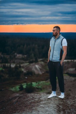 Tepede duran çekici genç adam ve ormanın manzarasına ve günbatımı bulutlu gökyüzüne bakıyor.