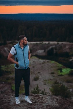 Tepede duran çekici genç adam ve ormanın manzarasına ve günbatımı bulutlu gökyüzüne bakıyor.