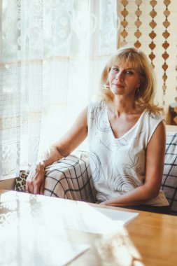 Zarif sarışın beyaz kadın portresi beyaz tişörtlü şık saçlı yaşlı kadın pencerenin yanındaki yemek masasında oturmuş neşeli bir gülümsemeyle kameraya bakıyor. güneşli hava.