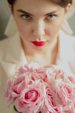 Güzel, mutlu, siyah saçlı, ceketinde beyaz bir fiyonk olan, kameraya bakan ve elinde bir buket gül tutan bir gelinin yakın çekimi. Kırmızı dudaklar