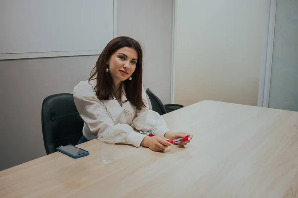 Mulher morena jovem em uma blusa branca sentada à mesa no escritório e segurando um marcador em suas mãos.. — Fotografia de Stock