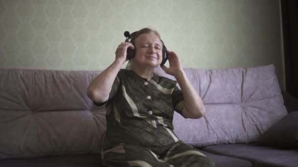 Babička ve sluchátkách, v dobré náladě poslouchá zábavnou hudbu. Babička ve sluchátkách poslouchá hudbu na gauči a tancuje. — Stock video