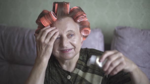 De oudere vrouw in de krullen doet de crème op haar gezicht. — Stockvideo