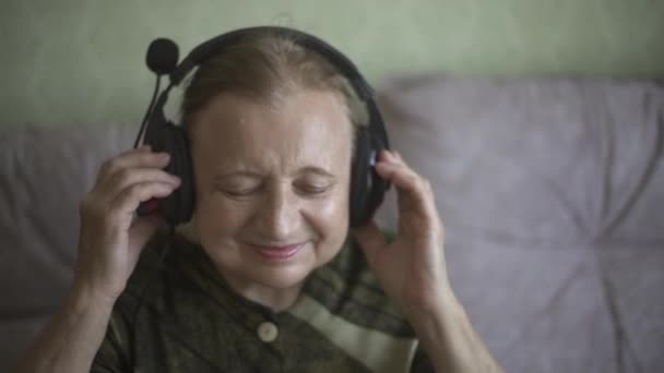 Grand-mère dans les écouteurs, d'une humeur magnifique écoute de la musique joyeuse. Grand-mère au casque écoute de la musique sur le canapé et danse sur la mélodie. — Video