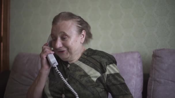 Nagymama izgatott, hogy hall valakit a telefonban, és vidáman beszél. Öregasszony kellemesen beszél a vezetékes telefonon ül a kanapén a nappaliban. — Stock videók