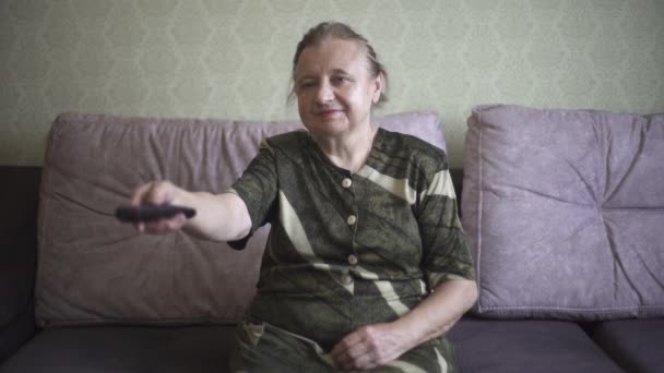 La abuela enciende felizmente un televisor. La anciana enciende un televisor y se sienta en el sofá. — Vídeo de stock