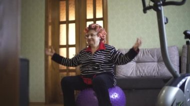 Spor giysili yaşlı bir kadın ve saçlarında bigudiler bir jimnastik topunun üzerinde oturuyor ve ellerini kaldırıyor. Spor kıyafetli büyükanne jimnastik topunda aerobik yapıyor..