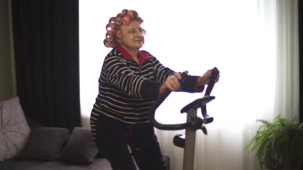 Starsza pani w odzieży sportowej i loki we włosach szczęśliwie pozbywa się na rowerze stacjonarnym i radośnie się uśmiecha. Babcia w odzieży sportowej robi aerobik na stacjonarnym rowerze.. — Wideo stockowe