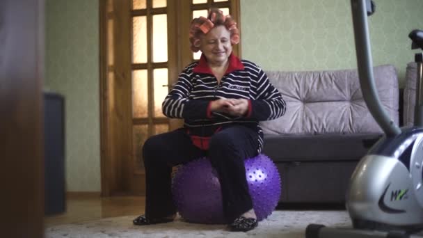 Die alte Dame in Sportkleidung und Lockenwickler im Haar sitzt auf einem Gymnastikball und macht Drehungen. Großmutter in Sportkleidung macht Aerobic auf einem Turnball. — Stockvideo