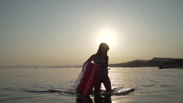 Молода дівчина виходить з моря з надувним матрацом в руках. Жінка виходить з вечірнього моря з надувним матрацом в руках . — стокове відео