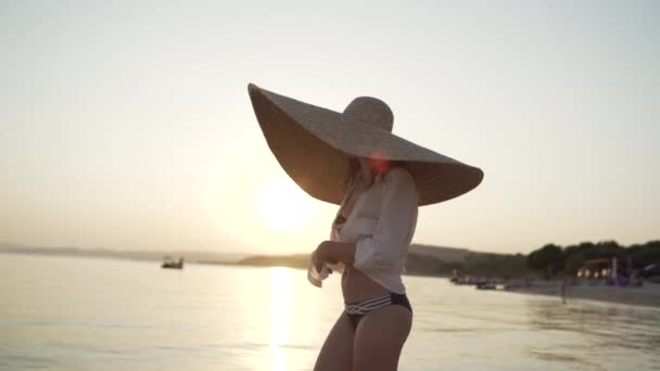 Tiro médio de menina com um chapéu grande andando perto do mar e amarrando sua camisa. Jovem mulher vestindo um chapéu grande e óculos de sol está andando na costa e amarrar a camisa. — Vídeo de Stock