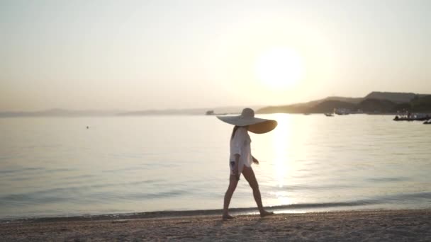 Tiro longo da menina em um chapéu grande que anda perto do mar. Jovem usando um chapéu grande está andando na costa e assistindo o pôr do sol. — Vídeo de Stock