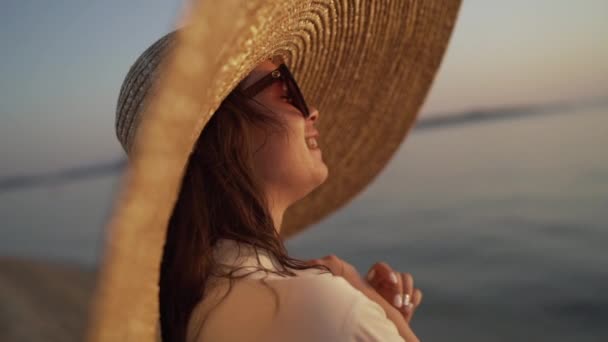 Retrato de close-up de uma menina usando um chapéu grande e assistindo o pôr do sol. Jovem senhora em um chapéu grande está sentado perto do mar e assistindo o pôr do sol. — Vídeo de Stock