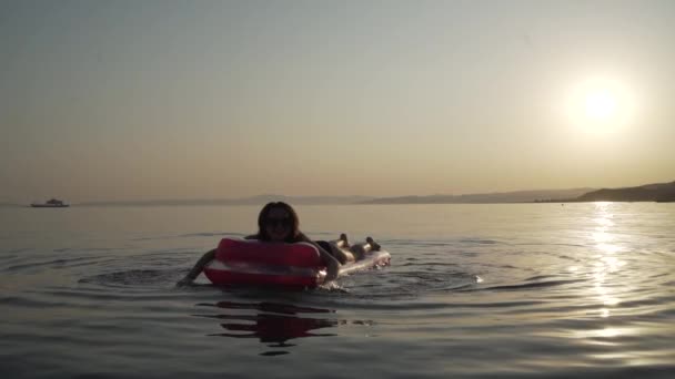 少女の中間ショット横たわって、夕方の海の膨脹可能なマットレスの上で泳いでいます。海のインフレータブルマットレスの上で横になって泳いでいる女性. — ストック動画