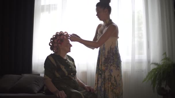 Νεαρή κοπέλα κάνει μαλλιά της γιαγιάς της. Νεαρή κυρία διαστρεβλώνει προσεκτικά τα μαλλιά της γριάς κυρίας σε ένα μπικίνι. — Αρχείο Βίντεο