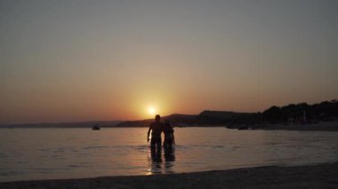 Çift, akşam denizinde gün batımını izlemekten mutlu. Kadın ve erkeğin uzun vadeli çekimi neşeyle denizde duruyor ve gün batımını seyrediyor..
