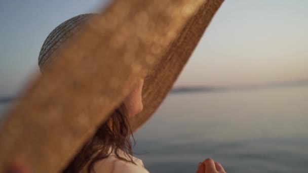 Büyük şapkalı, güneş gözlüklü ve gün batımını izleyen bir kızın yakın plan portresi. Büyük şapkalı genç bayan denizin kenarında oturuyor ve gün batımını seyrediyor.. — Stok video
