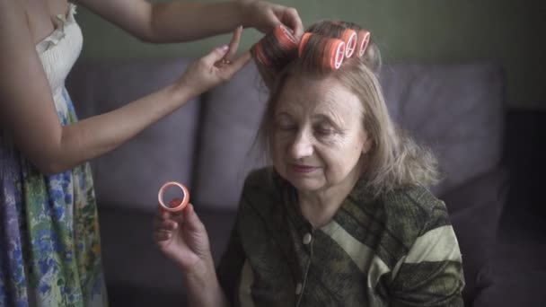 Νεαρή κοπέλα κάνει μαλλιά της γιαγιάς της. Νεαρή κυρία διαστρεβλώνει προσεκτικά τα μαλλιά της γριάς κυρίας σε ένα μπικίνι. — Αρχείο Βίντεο