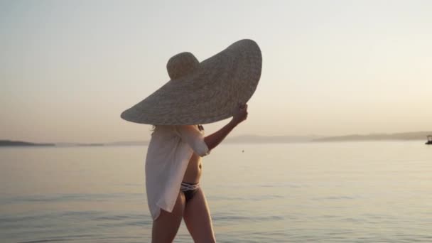 Tiro médio da menina em um chapéu grande que anda perto do mar e que gira sua cabeça para prestar atenção ao por do sol. Jovem mulher vestindo um chapéu grande e óculos de sol está andando na costa e assistindo o pôr do sol. — Vídeo de Stock