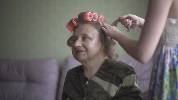 Młoda dziewczyna robi włosy swojej babci. Młoda dama bacznie przekręca włosy starszej pani na lokaju. — Wideo stockowe